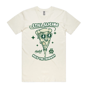An off-white short-sleeve t-shirt with a green graphic of a slice of pizza wearing sunglasses, with the text 'STILL SLICING' and 'HOLD THE FAIRWAY'.