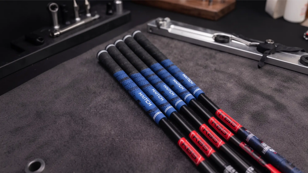 Six Haitch golf shafts arranged diagonally on a dark gray textured surface, showing black and blue handles with red and white Haitch branding.