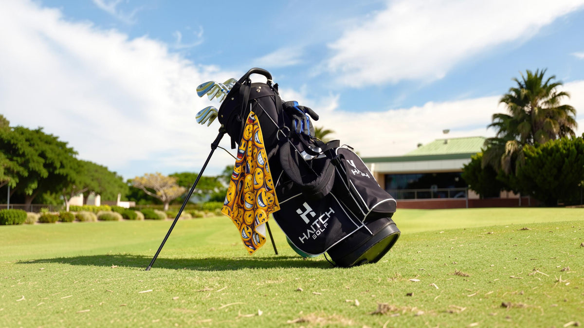 A black golf bag with white branding stands on a grassy golf course under a blue sky with clouds. A yellow and black patterned towel is draped over the bag, and golf clubs are visible inside.