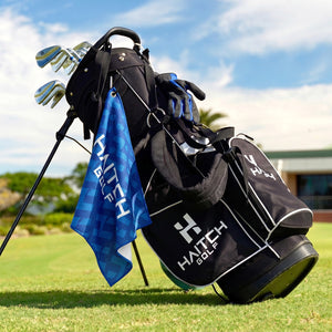 A black HAITCH GOLF stand bag filled with golf clubs and accessories, set on a grassy golf course under a blue sky. A blue patterned HAITCH golf flag is draped over the side of the bag.