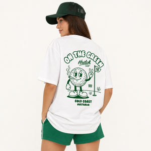 A woman wearing a white graphic t-shirt and green shorts, viewed from the back. The t-shirt features a green cartoon golf ball character with a flag and the text 'ON THE GREENS Haitch GOLF COLD COAST AUSTRALIA'.