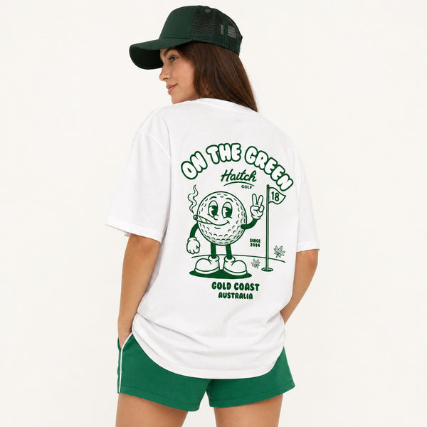 A woman wearing a white graphic t-shirt and green shorts, viewed from the back. The t-shirt features a green cartoon golf ball character with a flag and the text 'ON THE GREENS Haitch GOLF COLD COAST AUSTRALIA'.