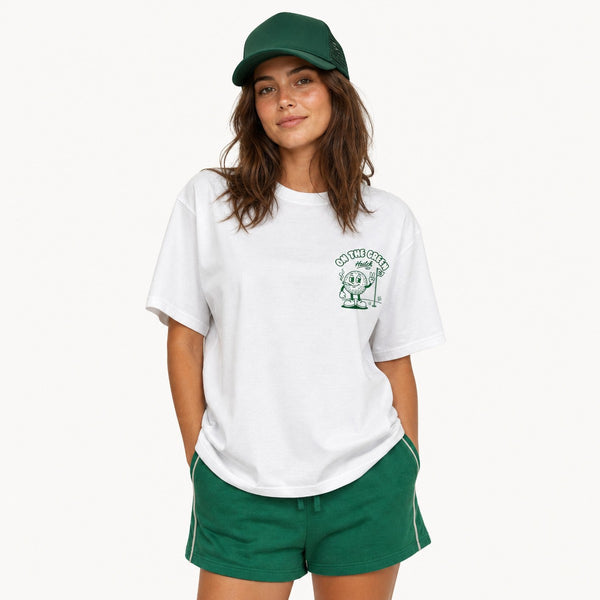 A woman modeling a white short-sleeve graphic t-shirt with a green chest print, paired with green shorts and a green cap, against a plain white background.