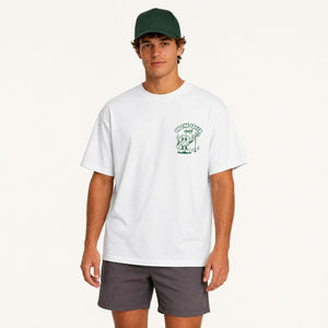 A man modeling a white crewneck t-shirt with a green chest graphic that reads 'ON THE GREEN' and features a cartoon character, paired with dark gray shorts and a dark green baseball cap, against a plain off-white background.