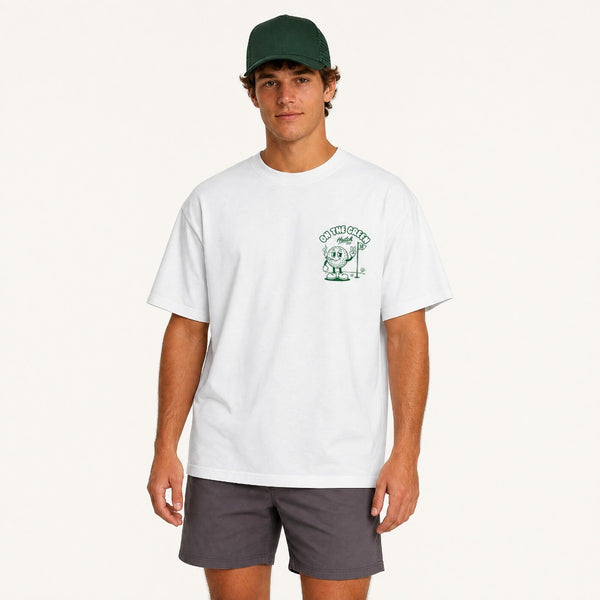 A man modeling a white crewneck t-shirt with a green chest graphic that reads 'ON THE GREEN' and features a cartoon character, paired with dark gray shorts and a dark green baseball cap, against a plain off-white background.