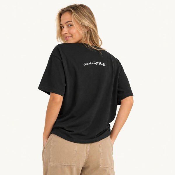 A woman wearing a black short-sleeve t-shirt with 'Smash Golf Balls' printed in white script on the back, paired with beige cargo pants, photographed against a clean off-white background.