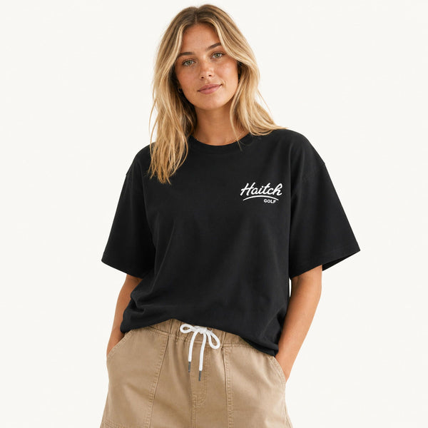 A woman wearing a black short-sleeve t-shirt with a white 'Haitch Golf' logo on the left chest, paired with beige drawstring pants, against a clean white background.