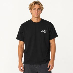 A man wearing a black short-sleeve crewneck t-shirt with a small white 'Haitch' logo on the left chest, paired with dark gray drawstring pants, against a plain white studio background.