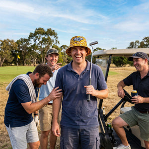 Four men laughing on a golf course near a golf cart. The central man wears a blue polo shirt, khaki shorts, and a yellow patterned bucket hat with 'Haitch Golf' printed on it.