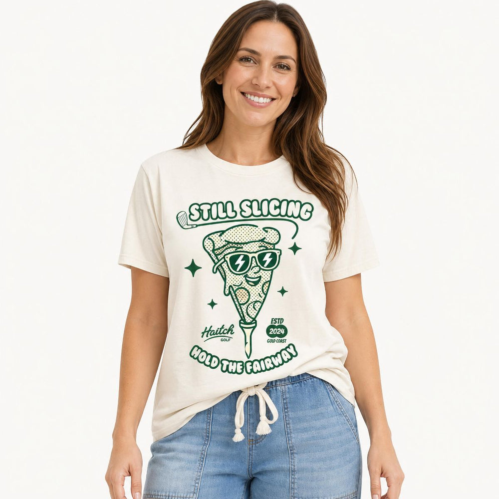 A woman wearing a cream-colored graphic t-shirt with a green pizza slice illustration and the text 'STILL SLICING HOLD THE FAIRWAY'. She is also wearing light blue denim jeans against a clean white studio background.