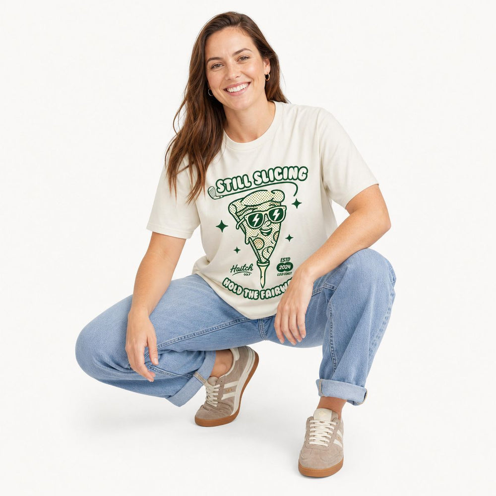 A woman wearing a cream-colored graphic t-shirt with a green pizza slice illustration and the text 'STILL SLICING HOLD THE FAIRWAY'. She is also wearing light blue denim jeans against a clean white studio background.