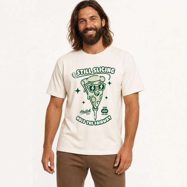 A man wearing a cream-colored t-shirt with a green graphic of a slice of pizza wearing sunglasses and the text 'STILL SLICING HOLD THE FAIRWAY'.