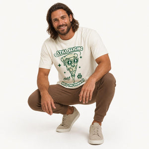 A man crouching in a studio setting wearing a cream-colored graphic t-shirt with a green pizza slice design and brown jogger pants.