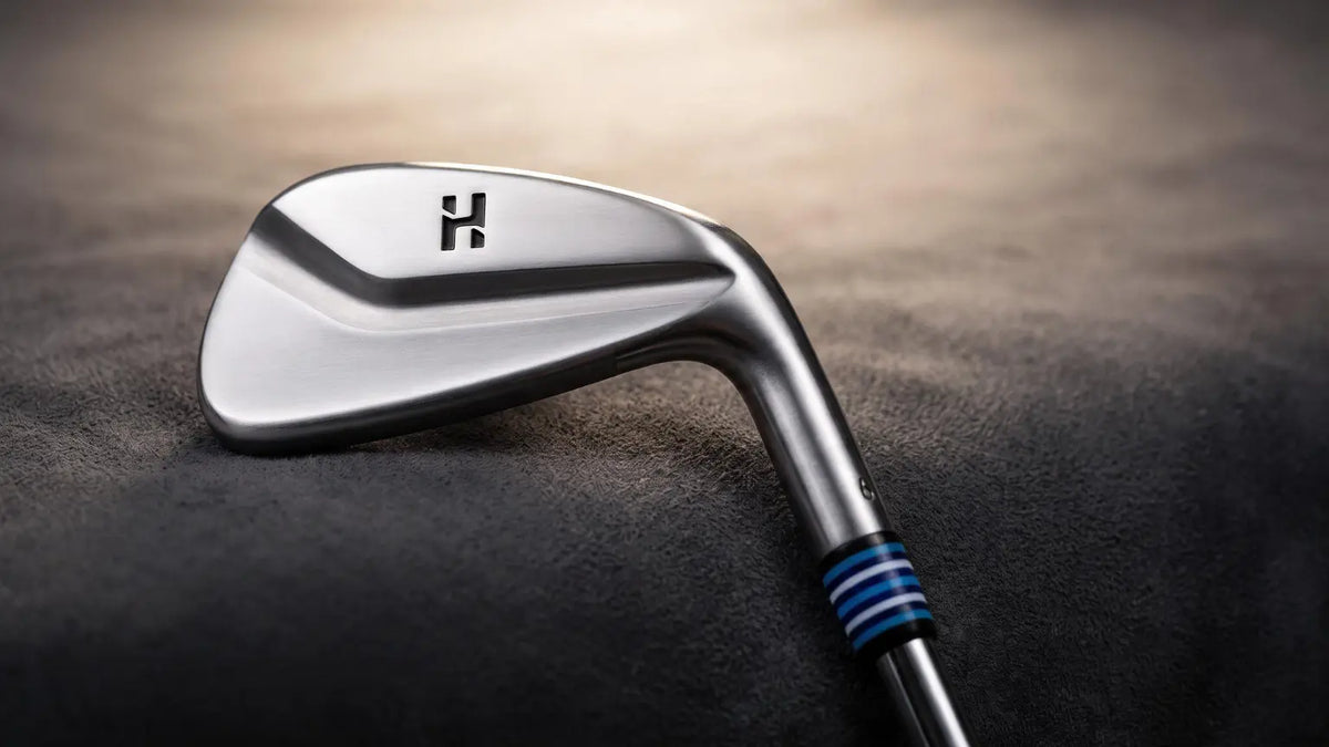 A close-up studio shot of a golf iron club head and part of the shaft resting on a dark textured surface. The club head is metallic silver with a black logo, and the shaft has blue and black grip stripes.
