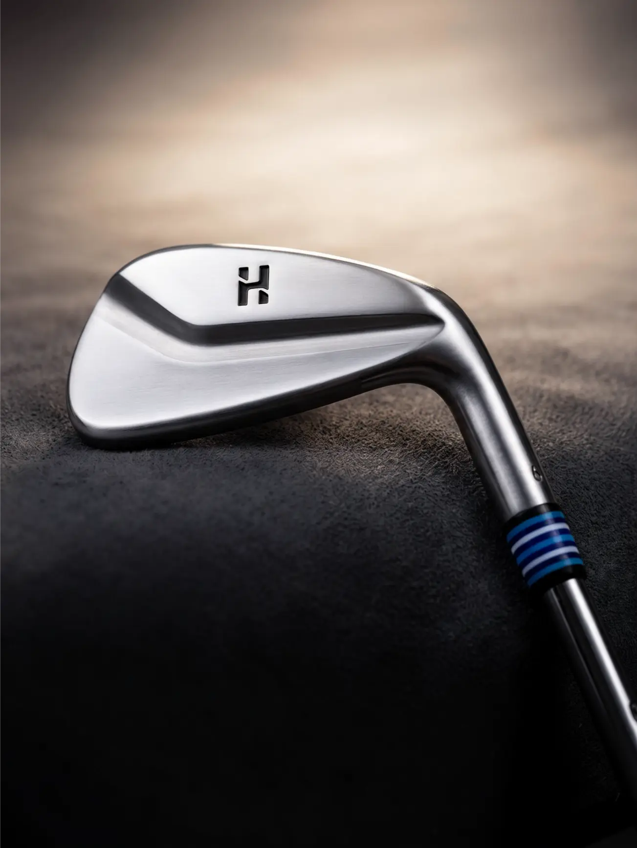 A close-up studio shot of a polished silver golf iron club head with a black logo and blue striped grip detail, resting on a dark textured surface with a warm blurred background.